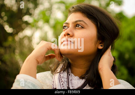 Ritratto di un simpatico paffuto viso tondo Indian ragazza adolescente guardando in alto e pensare, oltre bokeh di fondo la natura dello sfondo. Foto Stock