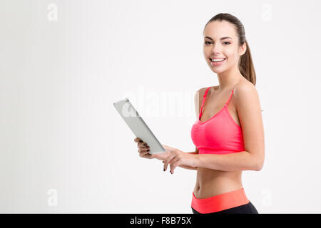 Positivo attraente giovane donna fitness in sportwear holding tablet su sfondo bianco Foto Stock