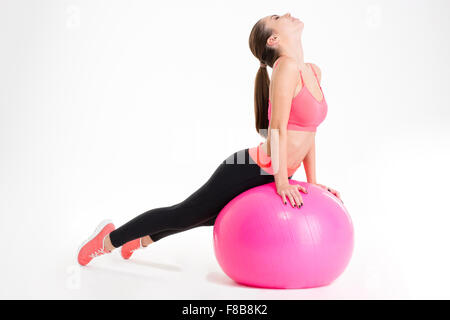 Focalizzato attraente giovane donna fitness in cima rosa e nero leggings facendo stretching su fitball rosa su sfondo bianco Foto Stock