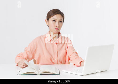 Donna adulta in maglietta arancione seduto alla scrivania che lavora sul computer portatile e in possesso di una pagina del libro con lo sguardo in avanti con la faccia aggrottò le sopracciglia Foto Stock