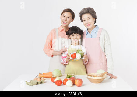 Giovane ragazza con una piastra con verdure in piedi tra la madre e la nonna dietro il tavolo con verdure e Foto Stock