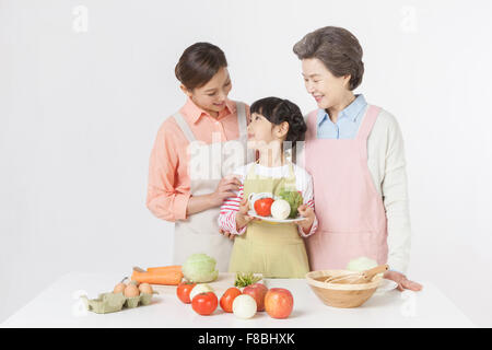 Giovane ragazza con una piastra con verdure in piedi tra la madre e la nonna dietro il tavolo con verdure e Foto Stock
