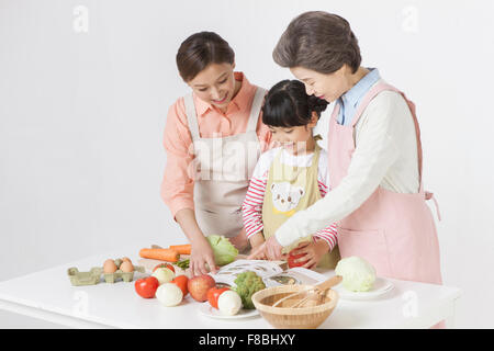 Figlia della madre e nonna di indossare un grembiule e guardando il libro di ricette sul tavolo con gli altri ingredienti e Foto Stock