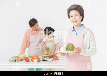Nonna tenendo una piastra con verdure con lo sfondo della madre e figlia cucina insieme Foto Stock
