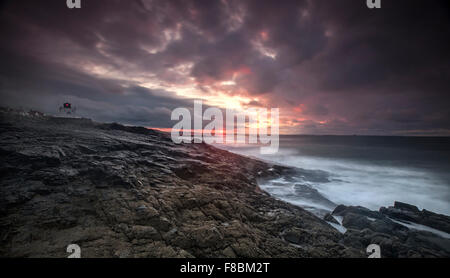 Il sole che tramonta la distanza al Trinity house stazione di luce sulle rocce stag a Bamburgh sulla costa di Northumberland Foto Stock