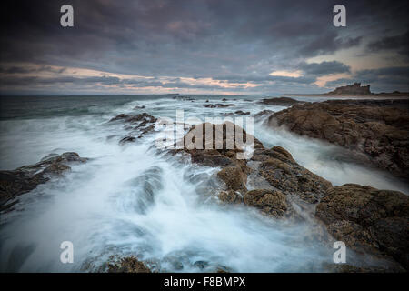 Una lunga esposizione delle onde che si infrangono sulle rocce di cervo al castello di Bamburgh sulla costa di Northumberland Foto Stock