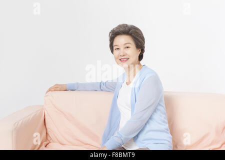 Senior donna con capelli corti in blu cardigan seduto su un divano e con lo sguardo in avanti con un sorriso Foto Stock