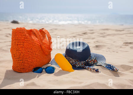 Borsa, cappello, crema solare e occhiali da sole sulla sabbia con lo sfondo della spiaggia al di fuori della messa a fuoco Foto Stock