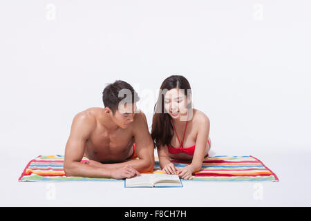 Matura in spiaggia indossare giacente sulla parte anteriore su una colorata asciugamano e guardando insieme un libro Foto Stock