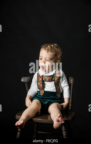 Un mese 18 old boy indossando lederhosen sbadigli mentre lui è seduta su una sedia in legno. Foto Stock