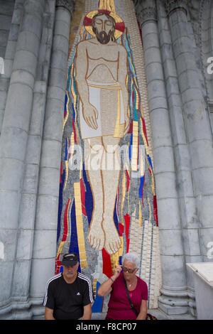 Giovane seduto davanti a un enorme scultura di Gesù in mosaico presso la chiesa principale di Loudes, Foto Stock