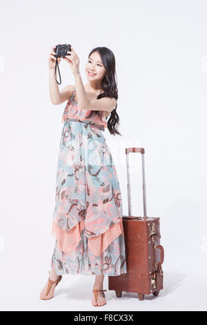 Donna con capelli lunghi in abito maxi e permanente tenendo una fotocamera con la sua valigia dietro di lei Foto Stock