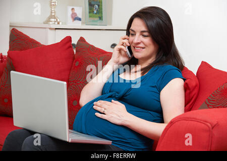 Donna incinta seduta sul divano con computer portatile e il telefono cellulare Foto Stock