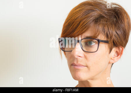 Vita ritratto di donna matura con i capelli rossi, occhi verdi, occhiali e grave espressione facciale, in piedi contro il wa Foto Stock