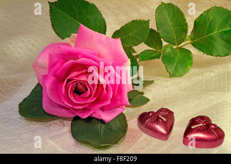 Un grande e luminoso giardino rosa rosa con foglie giacente su un bianco velo di seta. Prossimi due candele a forma di cuore. Foto Stock