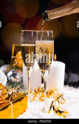 Versando bicchieri di champagne di festa da una bottiglia per festeggiare il Natale e il Nuovo Anno con candele accese e golden doni sulla neve invernale. Foto Stock