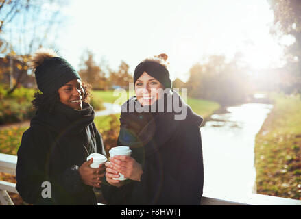 Due amiche guardando la fotocamera e sorridenti mentre si sta in piedi sul ponte, azienda di caffé, fiume sullo sfondo