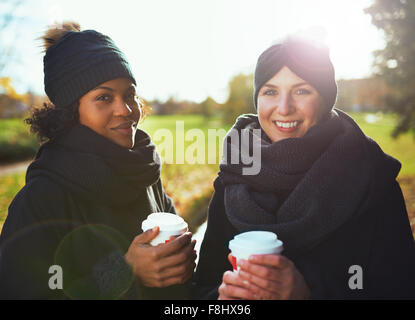 Due giovani donne guardando la telecamera, sorridenti mentre si tiene il caffè per andare contro di parco autunnali