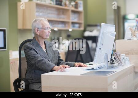 Senior addetta alla ricezione tramite computer desktop alla reception Foto Stock