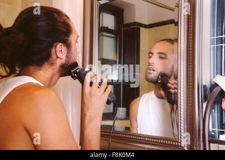 Giovane uomo guardando in uno specchio, la rasatura con il rasoio elettrico Foto Stock