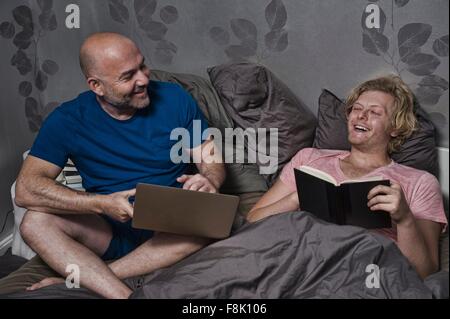 Voce maschile giovane seduto a letto a ridere mentre la lettura e utilizzo di computer portatile Foto Stock