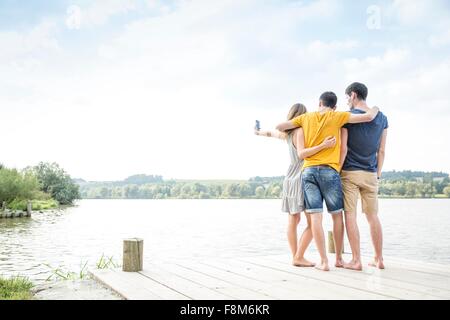Tre giovani adulti in piedi sul molo, tenendo autoritratto, utilizza lo smartphone, vista posteriore Foto Stock