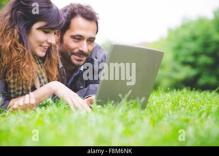 Testa e spalle di coppia giovane giacente sulla parte anteriore su erba utilizzando computer portatile sorridente Foto Stock
