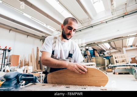 Giovane uomo nel laboratorio di falegnameria guardando verso il basso sullo skateboard di levigatura Foto Stock