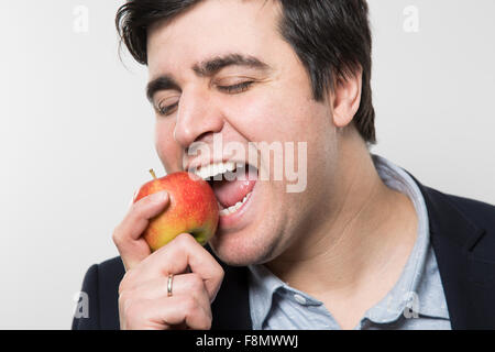 Dai capelli scuri imprenditore europeo con un look allegro prende un morso al di fuori di un piccolo apple con i suoi occhi chiusi mentre nella parte anteriore del Foto Stock