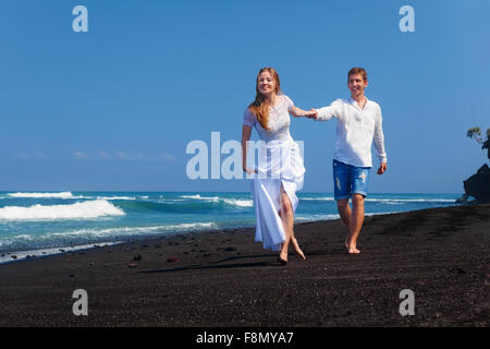 Felice famiglia sposi in luna di miele vacanze - appena sposato amare giovane eseguito con divertimento sul mare spiaggia di sabbia nera lungo il bordo dell'acqua. Foto Stock