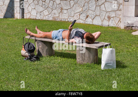 Conciate di mezza età donna caucasici in pantaloncini corti e occhiali da sole giacente dopo lo shopping su una panchetta outdoor e la lettura di libri elettronici Foto Stock