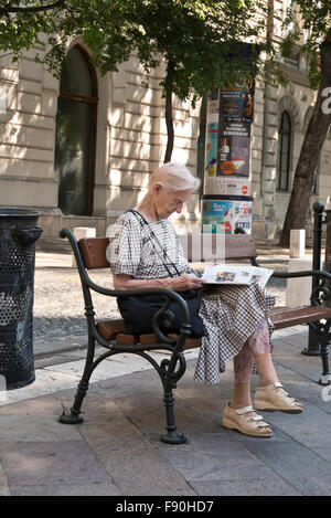 Un senior donna seduta su una panchina per strada e la lettura di un libro, Budapest, Ungheria. Foto Stock