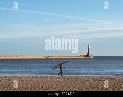 Uomo che porta driftwood lungo Roker beach con il North Pier e il faro dietro, Sunderland, England, Regno Unito Foto Stock