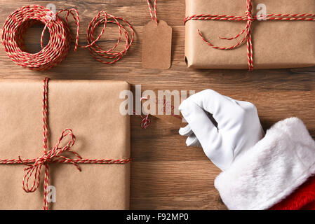 Angolo di alta vista di Santa Claus mano che tiene un regalo tag con buon Natale. Primo piano con regali impacchettati e string. Foto Stock