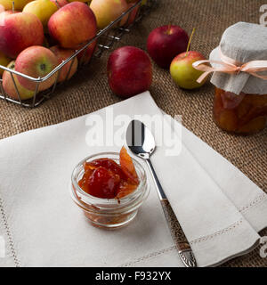 Il vasetto di vetro di marmellata di mele, cucchiaio, un vasetto di marmellata di arance, biancheria igienico e mele in filo metallico cesto su una tela coperti da tavola, square. Foto Stock