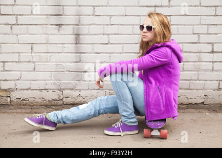 Bella bionda ragazza adolescente in occhiali da sole si siede su skateboard grigio vicino a un muro di mattoni Foto Stock