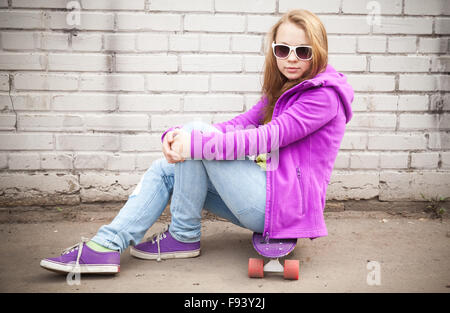 Bella bionda ragazza adolescente in occhiali da sole si siede su skateboard vicino al bianco muro di mattoni Foto Stock