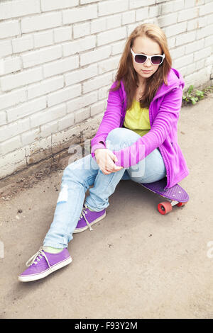 Bella ragazza adolescente in occhiali da sole si siede su skateboard urbano vicino a un muro di mattoni Foto Stock