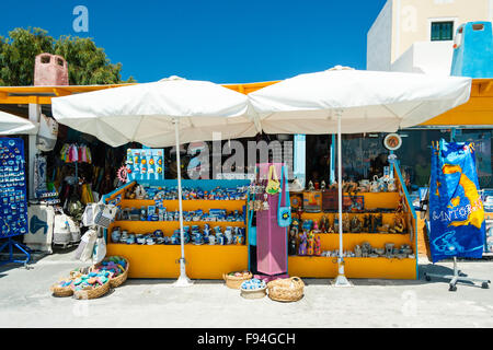 Santorini, Oia. Tipico turista negozio di souvenir con display esterno di due giallo e blu scaffalature in legno riempita con prodotti e due bianchi ombrelloni. Foto Stock
