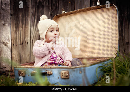 Felice bambina in vestiti caldi è seduta in valigia retrò di fronte la recinzione in autunno la natura Foto Stock