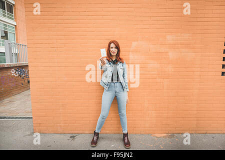 Giovani caucasici bello redhead capelli dritti donna posa appoggiata contro un muro di mattoni, mostrando una polaroid di se stessa, scattate con fotocamera istantanea - concetto di fotografia Foto Stock