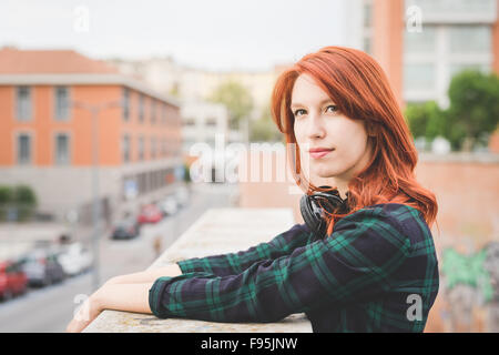 A metà lunghezza dei giovani caucasici bello redhead capelli dritti donna appoggiata su un corrimano, affacciato sulla sinistra, sereno, cuffie Foto Stock