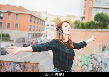 A metà lunghezza dei giovani caucasici bello redhead capelli dritti donna ascolto di musica con cuffie, sentendosi liberi con gli occhi chiusi e braccia aperte - libertà, musica, spensierata concept Foto Stock