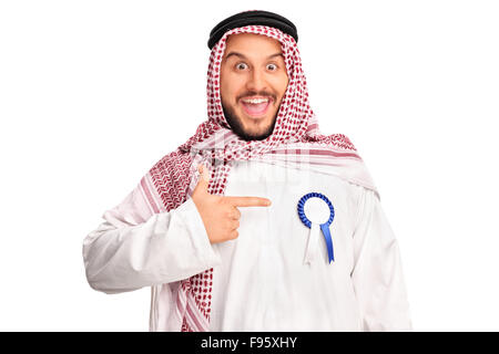 Gioiosa giovane arabo indossando un premio nastro sulla sua tunica e rivolti verso di essa con la sua mano isolati su sfondo bianco Foto Stock