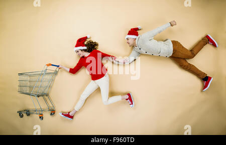 Coppia giovane di cappelli di Natale il divertimento in esecuzione con il carrello della spesa contro lo sfondo beige Foto Stock