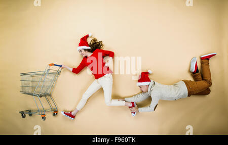 Coppia giovane di cappelli di Natale il divertimento in esecuzione con il carrello della spesa contro lo sfondo beige Foto Stock