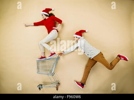 Coppia giovane di cappelli di Natale il divertimento in esecuzione con il carrello della spesa contro lo sfondo beige Foto Stock