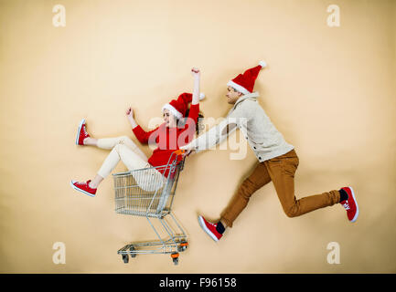 Coppia giovane di cappelli di Natale il divertimento in esecuzione con il carrello della spesa contro lo sfondo beige Foto Stock