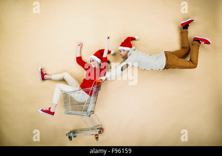 Coppia giovane di cappelli di Natale il divertimento in esecuzione con il carrello della spesa contro lo sfondo beige Foto Stock