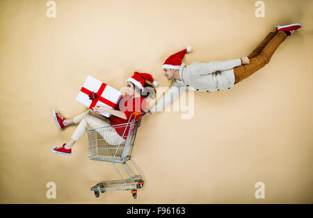 Coppia giovane di cappelli di Natale il divertimento in esecuzione con il carrello della spesa contro lo sfondo beige Foto Stock